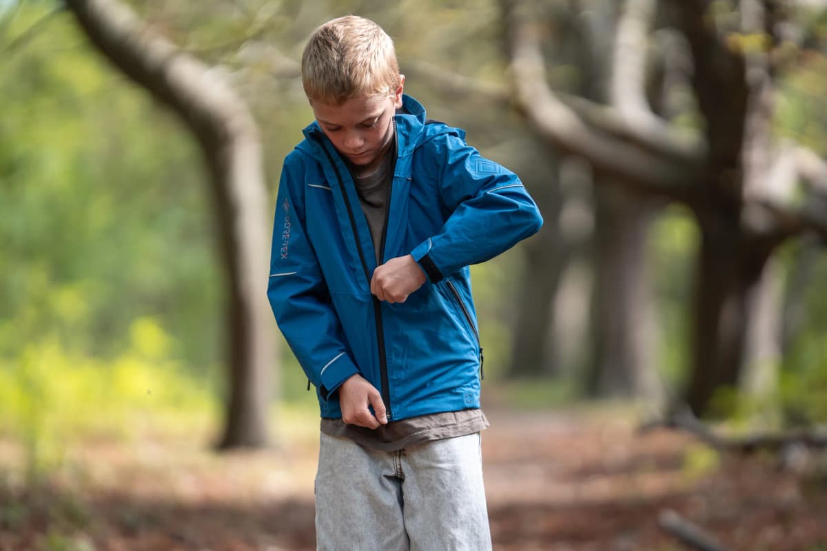 A boy zipping up a blue GORE-TEX Kidswear jacket in a tree-lined avenue