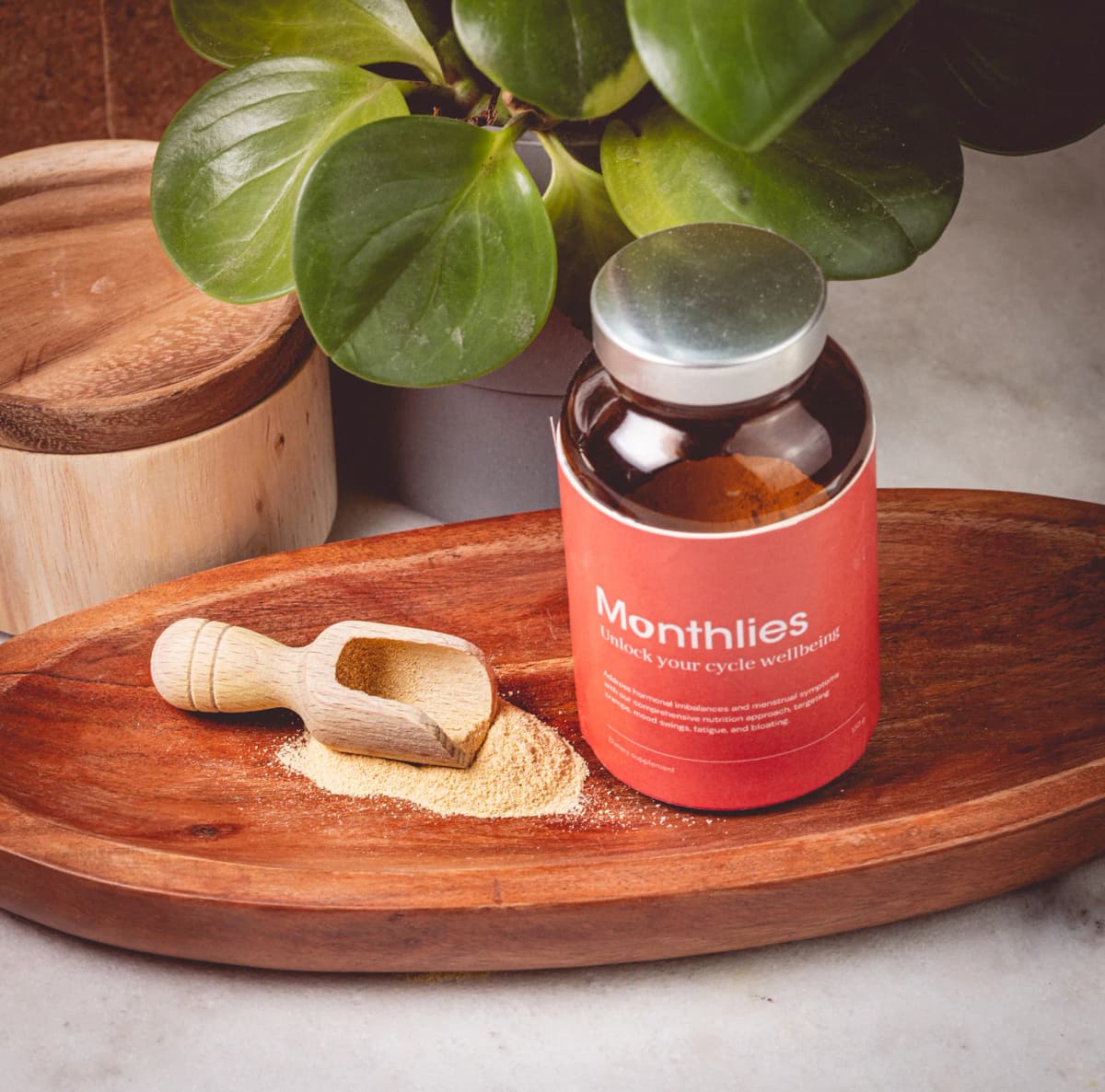 The Monthlies jar on a wooden tray beside a scoop of powder and a plant