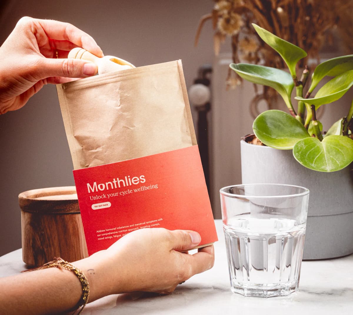 Hands opening the Monthlies refill pouch beside a glass of water and a plant